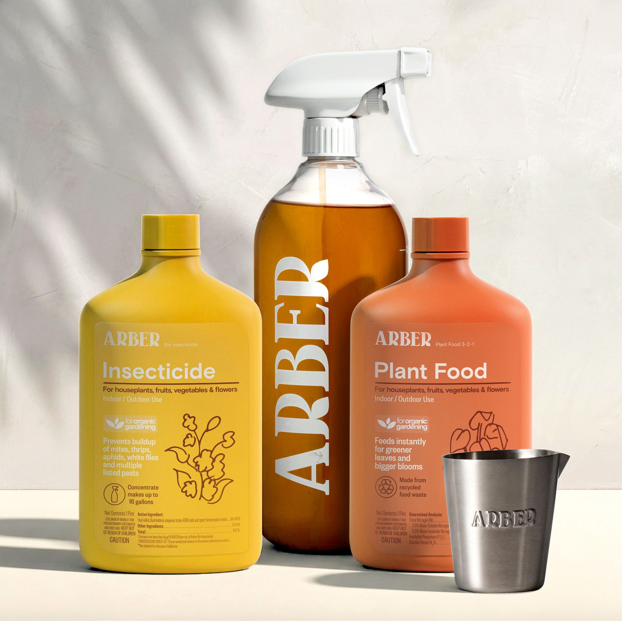 Three gardening product bottles and a metal measuring cup arranged on a white surface with shadows